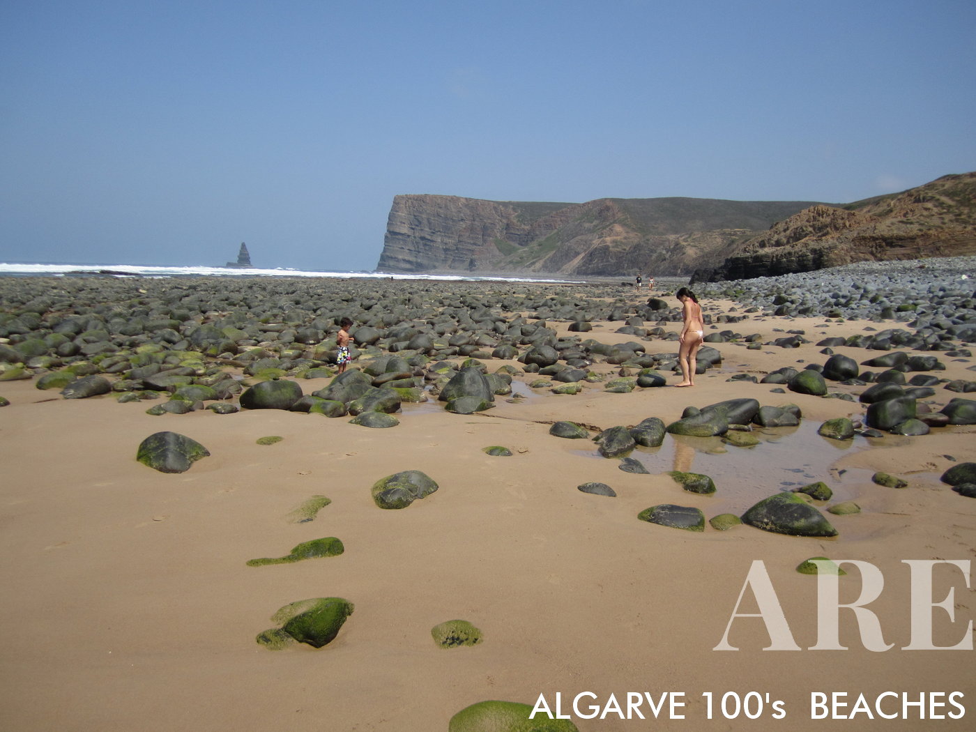 Canal Beach en Aljezur, Algarve, presenta un panorama encantador de pequeñas rocas, olas rítmicas y el vasto océano, que culmina en la silueta distintiva de la roca Ponta da Agulha en el horizonte.
